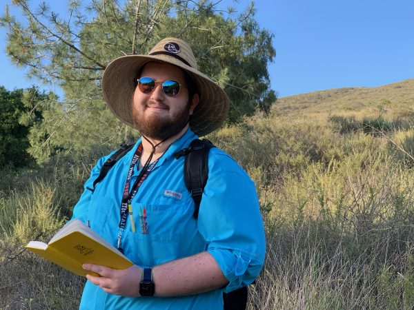 Liam Schramm standing in a grassy field holding a yellow notebook wearing a full brimmed hat, sun glasses and a blue shirt