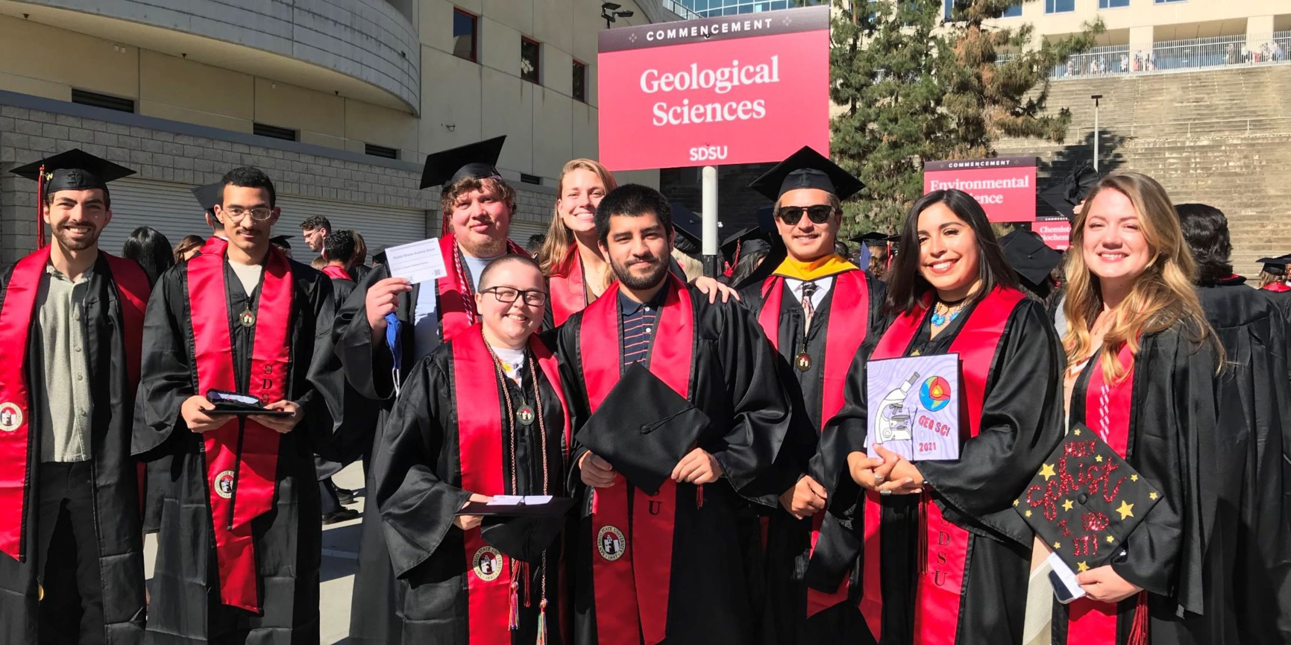 Undergraduates and graduates wearing graduation regalia gathered together in front of the Geological Sciences sign